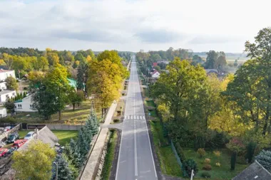 Widok z lotu ptaka na proste, dwupasmowe drogę wiodącą przez zabudowaną wieś, otoczoną drzewami w jesiennych barwach.